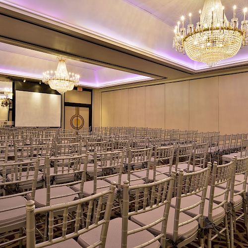 A conference room with rows of chairs, chandeliers, and a projector screen, prepared for an event or lecture, with elegant lighting and decor.