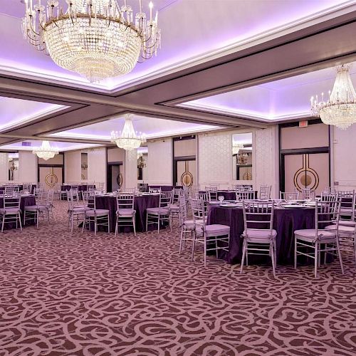 The image shows an elegant banquet hall with chandeliers, round tables draped in purple cloths, silver chairs, and patterned carpet, set for an event.