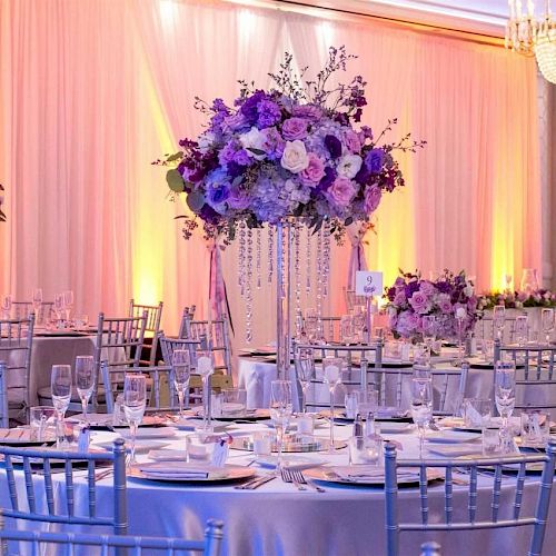 Elegant event setup with lavender and white floral arrangements, round tables, and silver chairs. Chandeliers and warm lighting create ambiance.