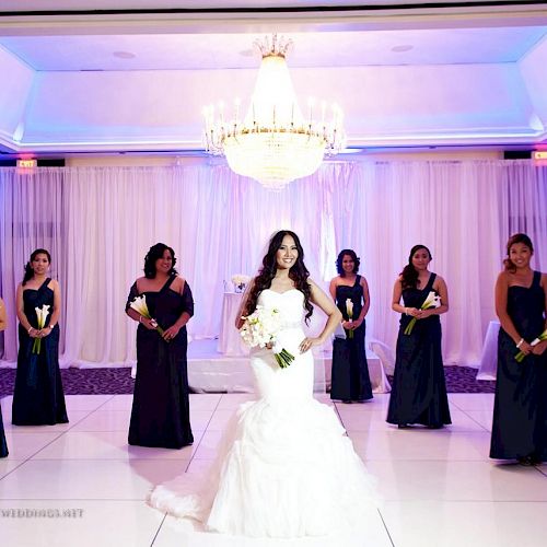 A bride in a white gown stands with six bridesmaids in dark blue dresses, each holding flowers, under a chandelier in a decorated venue.