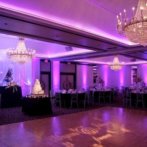 An elegant ballroom decorated for a wedding with chandeliers, purple lighting, and a wedding cake centerpiece.