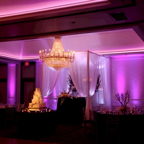 A dimly lit ballroom decorated with purple lighting, featuring a grand chandelier, draped white curtains, a wedding cake, and elegant table settings.