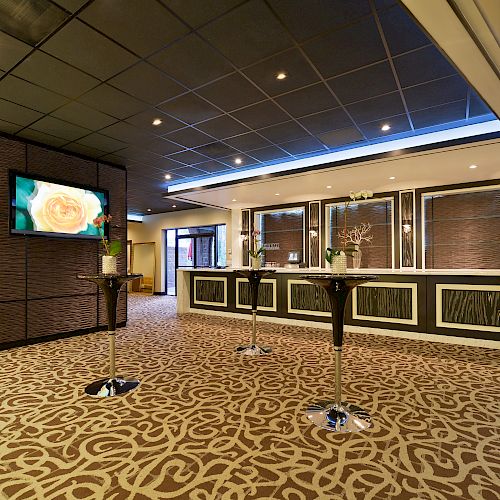 The image shows a modern hotel lobby with a reception desk, patterned carpet, barstool tables, and television screens displaying an image.