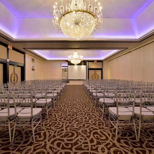 The image shows a conference or event hall setup with rows of silver chairs, chandeliers, and a large projector screen at the front of the room.