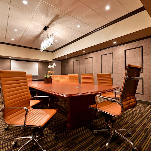 A modern conference room with a wooden table, six orange chairs, a projector screen, and a decorative plant on the table.