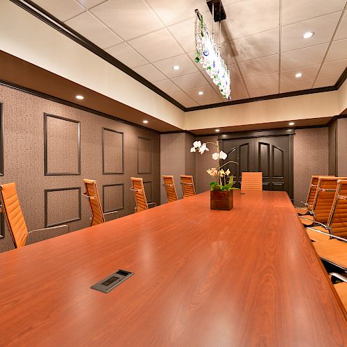 A modern conference room with a long wooden table, several brown chairs, and a plant centerpiece under a ceiling light.