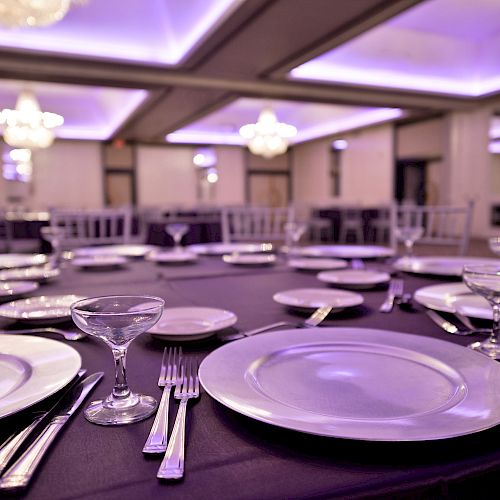 A formal dining setup with plates, utensils, and glassware on a round table, in an elegant room with chandeliers and purple ambient lighting.