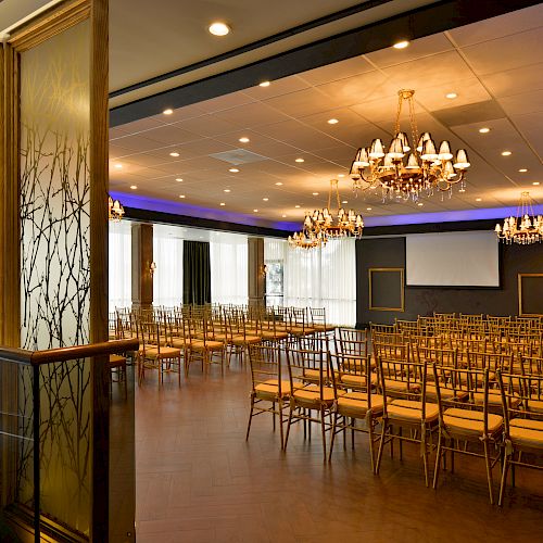 A well-lit, elegant conference room with rows of gold chairs, chandeliers, a projection screen, and a decorative room divider.