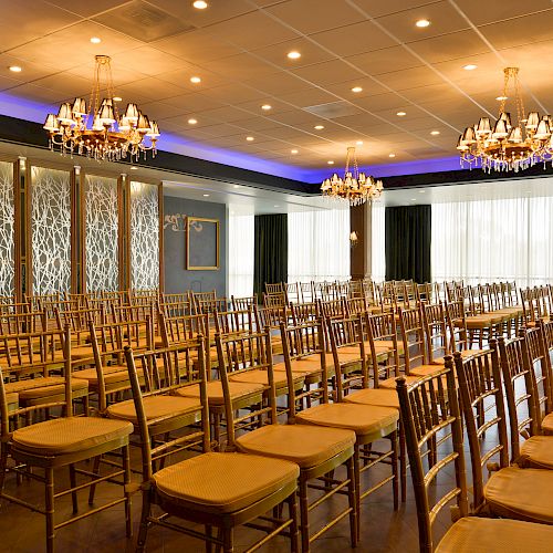 A well-lit, elegant room with rows of chairs arranged auditorium-style, chandeliers overhead, and decorative partitions, possibly set up for an event.