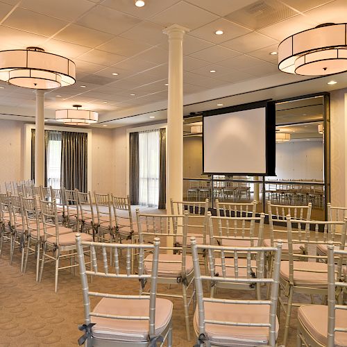 The image shows a conference room with rows of chairs facing a screen, soft lighting, and large windows with curtains.