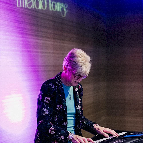 A person is playing an electronic keyboard on stage with a sign in the background that reads "Emerald Lounge". The lighting is purple and blue.