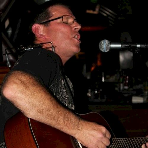 A person is playing an acoustic guitar and singing into a microphone, wearing glasses and appearing to be in a performance setting, with closed eyes.