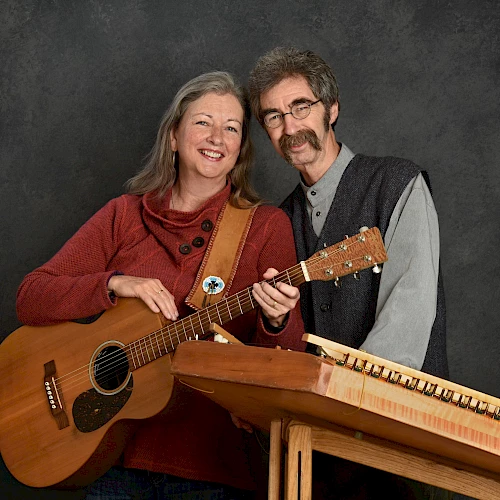 A smiling woman holding a guitar and a man with a stringed instrument, posing in front of a dark background.