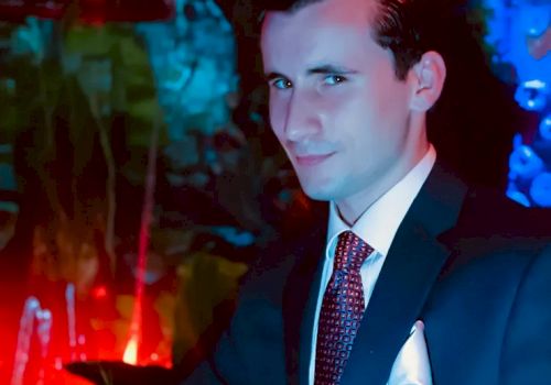 A man in a suit and tie poses indoors with colorful, dim lighting in the background, creating a somewhat mysterious or nightclub-like atmosphere.