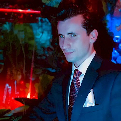 A man in a suit and tie poses indoors with colorful, dim lighting in the background, creating a somewhat mysterious or nightclub-like atmosphere.