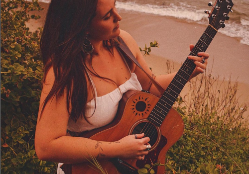 A person in a white dress is playing an acoustic guitar near a beach, standing among greenery with the ocean in the background.