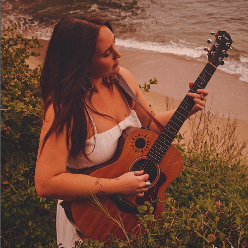 A person in a white dress is playing an acoustic guitar near a beach, standing among greenery with the ocean in the background.