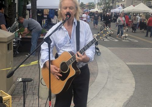 A person is performing with an acoustic guitar and microphone setup on a street with booths and tents in the background, possibly a market.