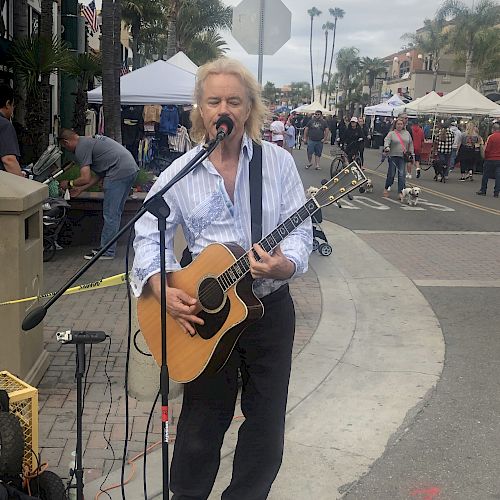 A person is performing with an acoustic guitar and microphone setup on a street with booths and tents in the background, possibly a market.