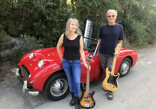 A woman and a man stand in front of a red vintage car, each holding musical instruments: a bass guitar and an electric guitar.