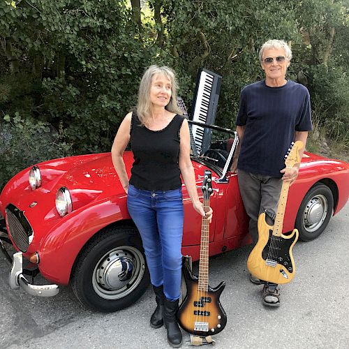 A woman and a man stand in front of a red vintage car, each holding musical instruments: a bass guitar and an electric guitar.