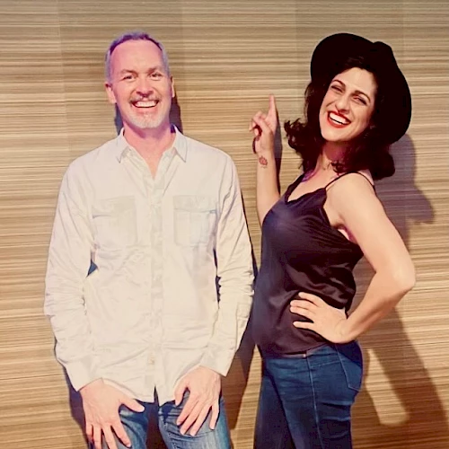 Two people are posing and smiling in front of a sign that says "Emerald Lounge," one standing casually and the other pointing at the sign.