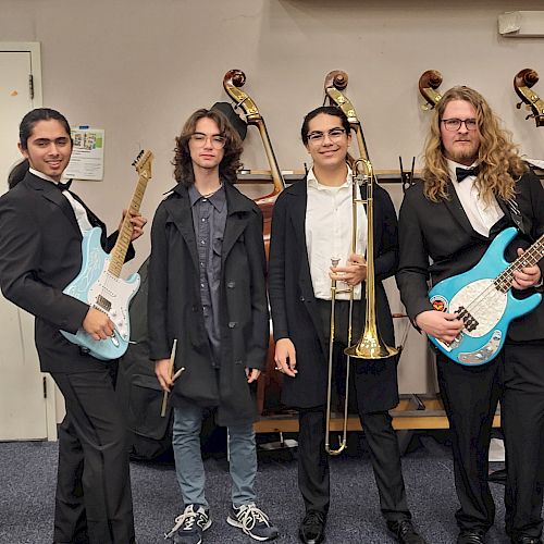 The image shows four people standing in front of a shelf with trophies. One holds a guitar, one a trombone, and two wear formal attire with guitars.