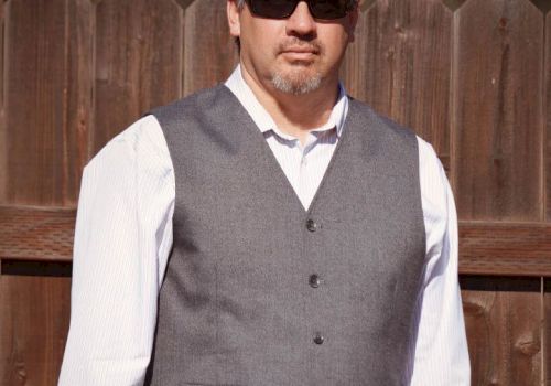 A man is standing in front of a wooden fence, wearing a white shirt, gray vest, and dark sunglasses.