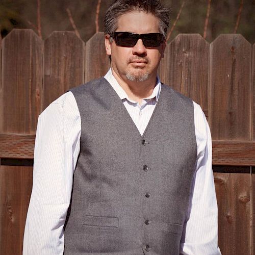 A man is standing in front of a wooden fence, wearing a white shirt, gray vest, and dark sunglasses.