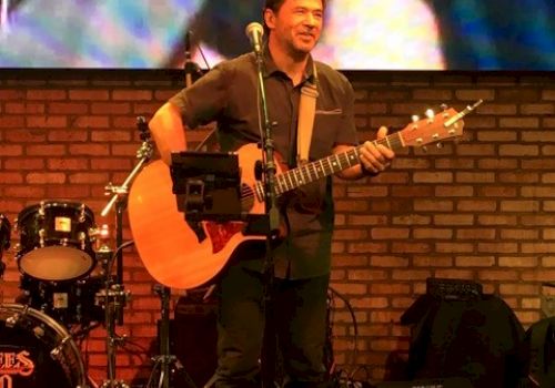 A musician is performing on stage with an acoustic guitar. There are drums in the background and a large screen displaying visuals behind him.