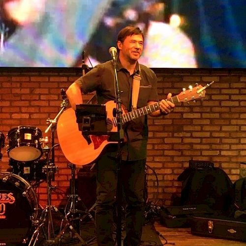 A musician is performing on stage with an acoustic guitar. There are drums in the background and a large screen displaying visuals behind him.