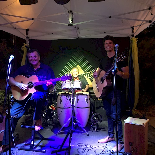 Three musicians are performing on a stage: two with guitars, one with drums. They are under a tent with microphones and stage lighting.