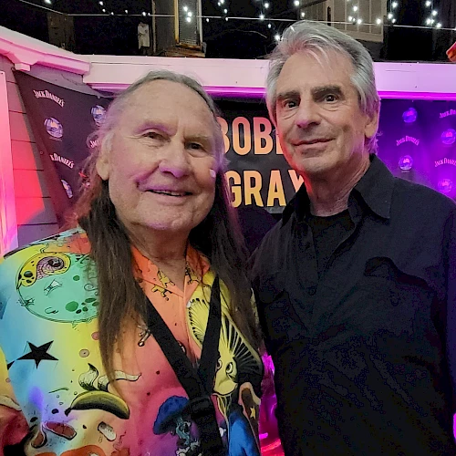Two men are standing together, smiling at an indoor event with colorful lighting and a sign in the background that reads "ROB" with other letters obscured.
