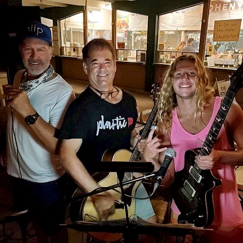 Three people posing with guitars in front of a café or restaurant, smiling. The background features lit windows with various posters.