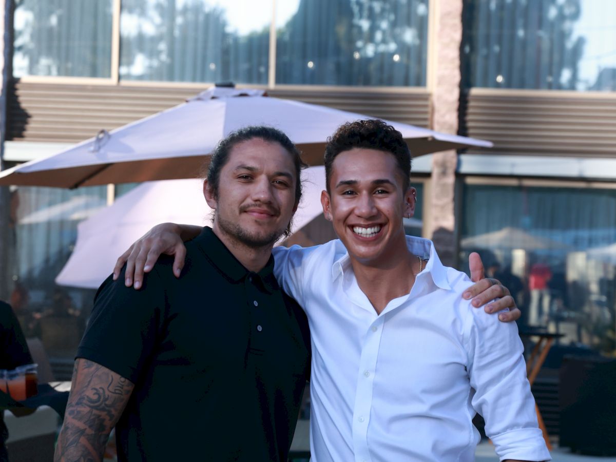 Two men are smiling and posing together outdoors in front of a building. One has a tattoo on his arm, and they're standing arm in arm.