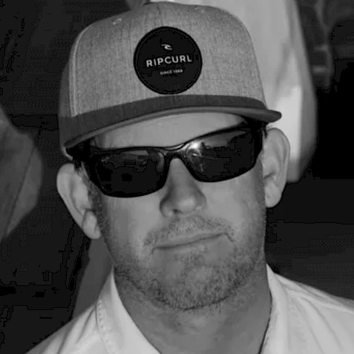 An individual wearing a "Rip Curl" cap, sunglasses, and a collared shirt, in a grayscale photo.
