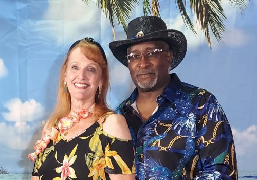 A smiling woman and man in tropical-themed attire pose in front of a backdrop with palm trees and a blue sky.