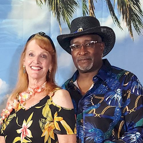 A smiling woman and man in tropical-themed attire pose in front of a backdrop with palm trees and a blue sky.