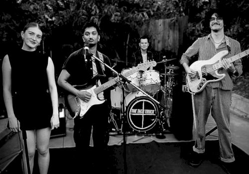 A four-member band is performing outdoors, featuring a drummer, two guitarists, and a vocalist, with trees in the background.