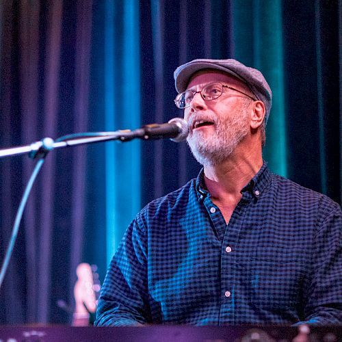 A man is singing and playing a keyboard on stage, wearing a flat cap, glasses, and a checked shirt, with colorful stage lighting in the background.