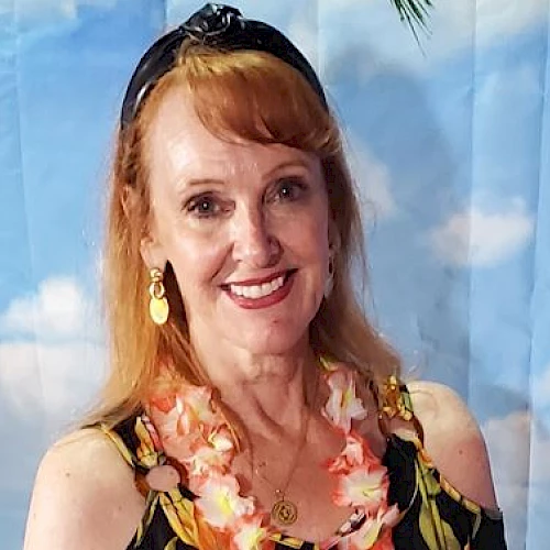 A smiling woman with red hair wears a floral lei and a black headband, posing in front of a backdrop with clouds, suggesting a tropical theme.