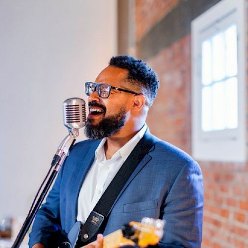 A man wearing a suit is singing into a microphone while playing an electric guitar. He has glasses and a beard, and the background shows a brick wall.