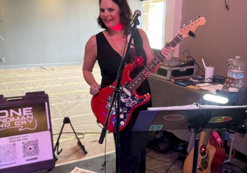 A woman with a red guitar is smiling and standing on a rug. There’s music equipment around her in a room with a partly visible large window.