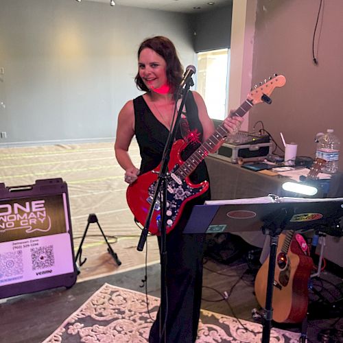 A woman with a red guitar is smiling and standing on a rug. There’s music equipment around her in a room with a partly visible large window.