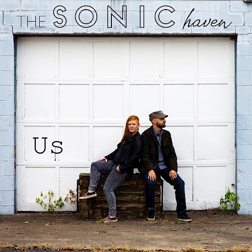 Two people sit on a wooden crate in front of a garage door with "THE SONIC haven" and "Us" written on it. They appear relaxed and casual.