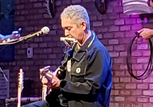 A person is playing a guitar and harmonica simultaneously on stage in front of a brick wall, while another person holds a cable in the background.