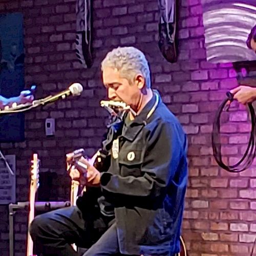 A person is playing a guitar and harmonica simultaneously on stage in front of a brick wall, while another person holds a cable in the background.