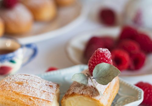 A plate with a cream-filled pastry topped with raspberries sits in focus, surrounded by additional raspberries and pastries in the background.