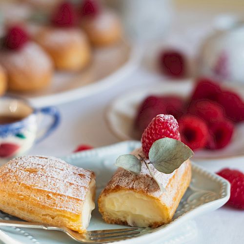 A plate with a cream-filled pastry topped with raspberries sits in focus, surrounded by additional raspberries and pastries in the background.