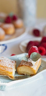 The image shows a plate with cream-filled pastries dusted with powdered sugar, garnished with raspberries and leaves. More pastries and raspberries are in the background.